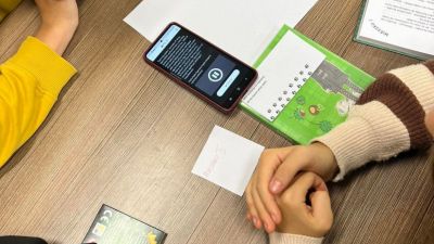 Troje dzieci korzystające z telefonu z otwartą aplikacją Actionbound. Na drugim planie materiały do wykorzystania w grze.
