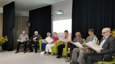 Klub Seniora "As" prezentujący fragment "Moralność Pani Dulskiej" - siedzą w zielonych fotelach, w tle niebieska kurtyna