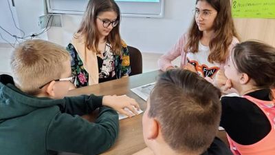 5 osób z klasy 6 rozwiązująca zadanie związane z twórczością M. Konopnickiej. W tle tablica interaktywna i postać Marii Konopnickiej.